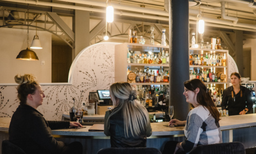 Three people sitting  at the bar drinking wine at Heist Restaurant.