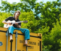 Nolan Hubbard sitting on top of a yellow shipping container, playing his guitar with lush trees in the background.