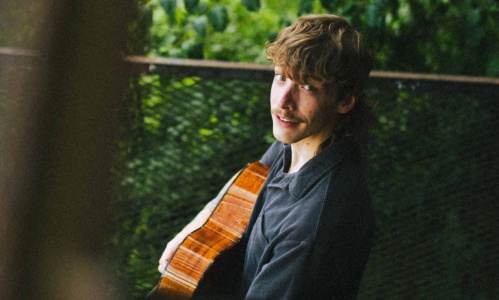 Nolan Hubbard playing his guitar while sitting down in front of green foliage.