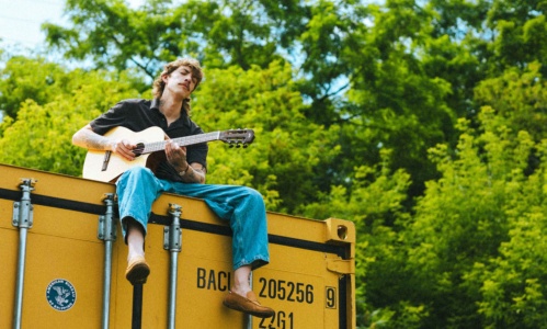 Nolan Hubbard sitting on top of a yellow shipping container, playing his guitar with lush trees in the background.