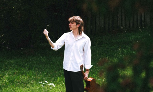 Nolan Hubbard holding his guitar and a small flower as he stands on lush green grass.